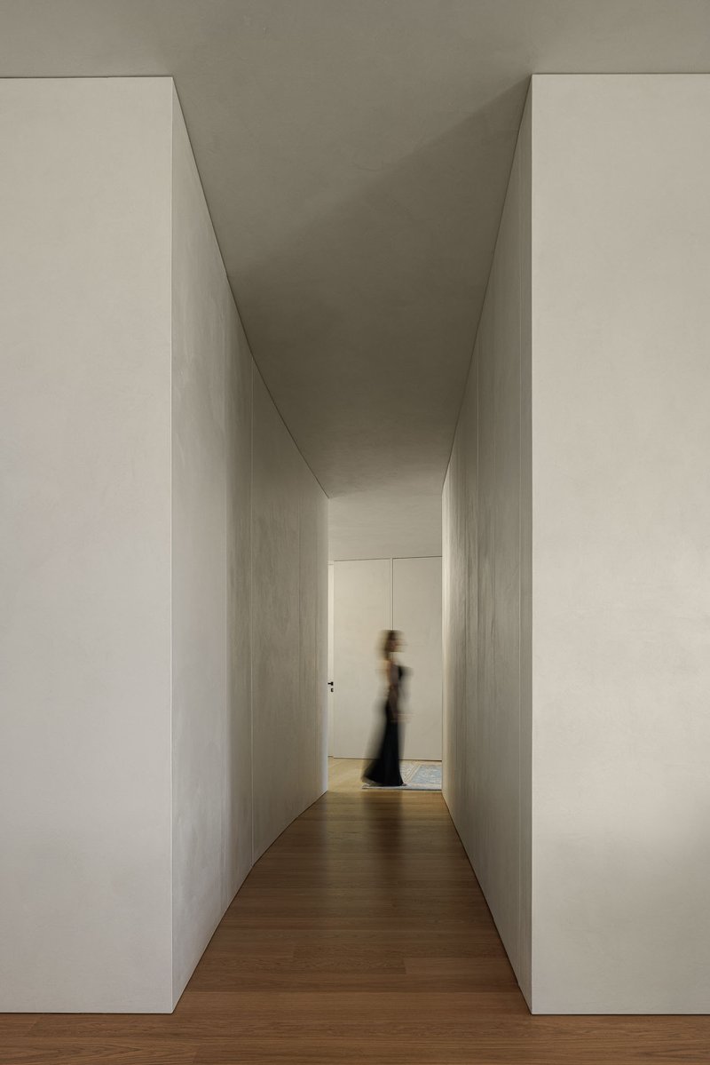 Narrow corridor with slanted concrete walls and timber floor leading toward a figure in shadow