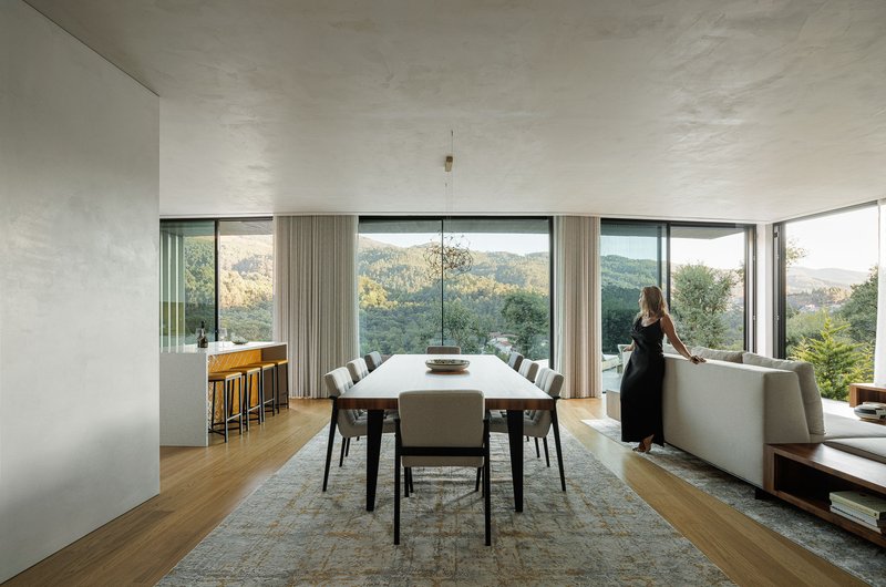 Open-plan dining area with continuous glass walls framing forested hills and a person at window