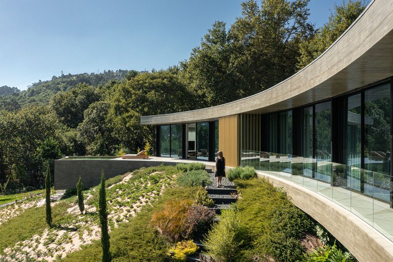 Curved concrete roof over glazed facade and planted green roof terrace with a person standing