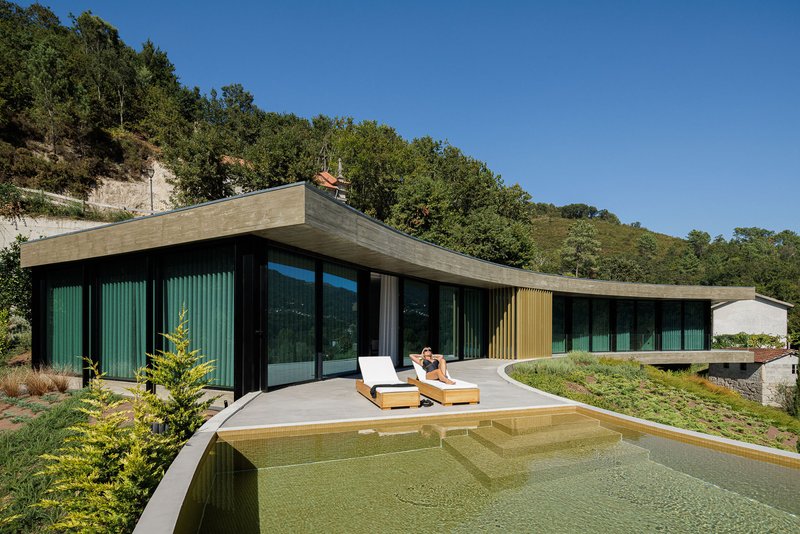 Cantilevered concrete roof above glass-walled pavilion and swimming pool with lounge chairs under clear sky