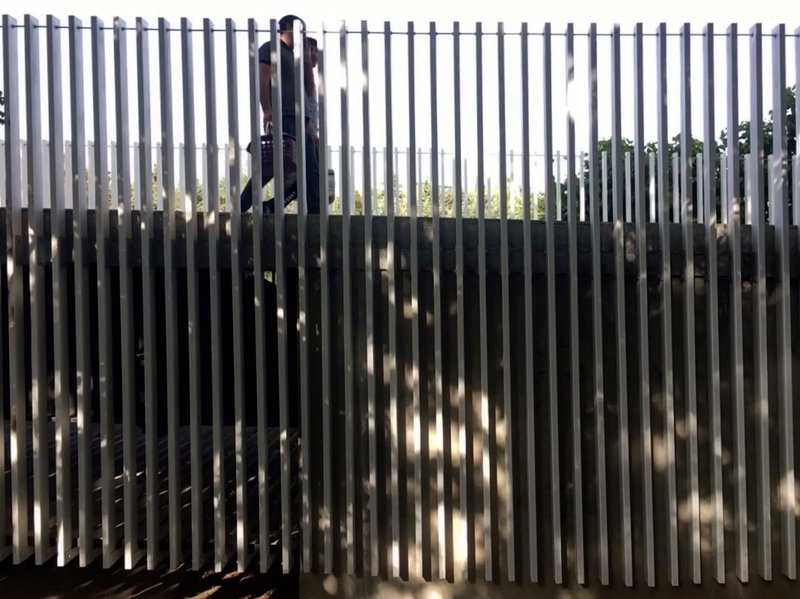 Figure walking behind vertical metal slats creating alternating bands of light and shadow