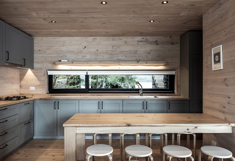 Kitchen with wood plank walls, grey cabinetry and a long horizontal window overlooking lakeside trees