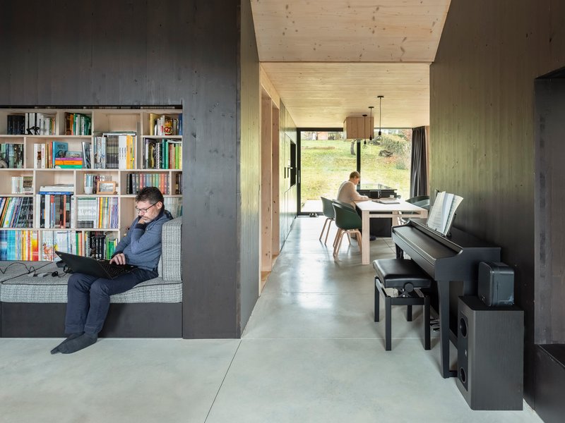 Interior view through concrete walls showing a man reading by the bookshelf and a woman at the desk