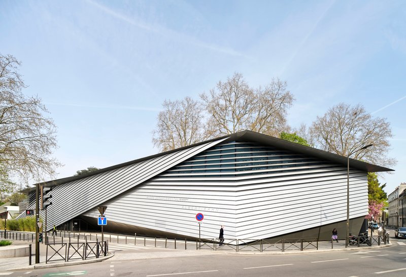 Street view of the sculptural ribbed metal facade with bare trees in the background