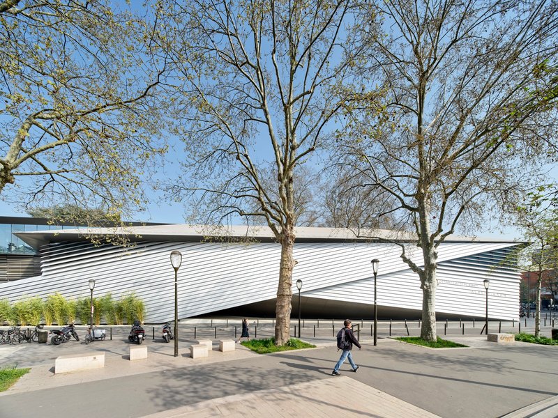 Horizontal metal louvers wrapping the curved facade as pedestrians walk beneath spring trees