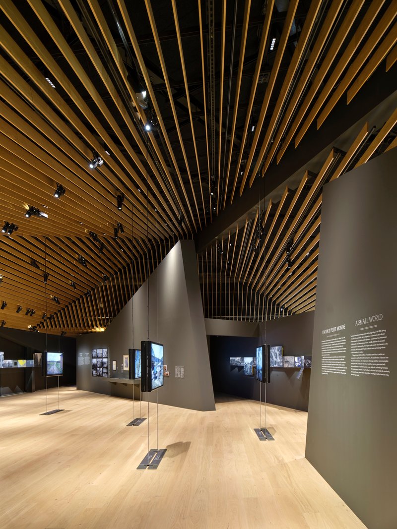 Exhibition gallery with radiating timber baffle ceiling and grey partition walls displaying photographs and text panels