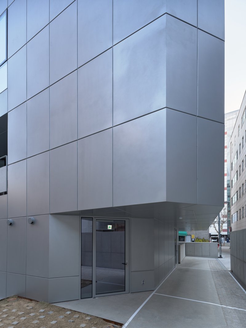 Exterior corner with pale grey metal panel facade, recessed glass entrance and covered walkway along the sidewalk