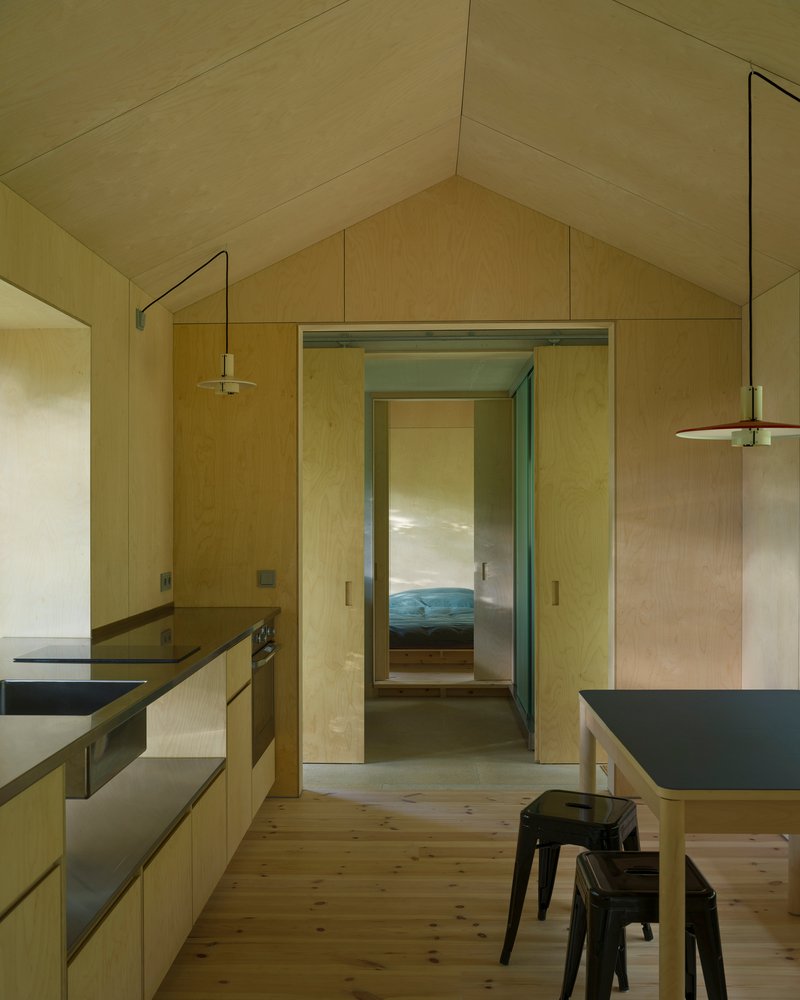 Plywood-lined kitchen with black countertops and view through doorway to bedroom beyond