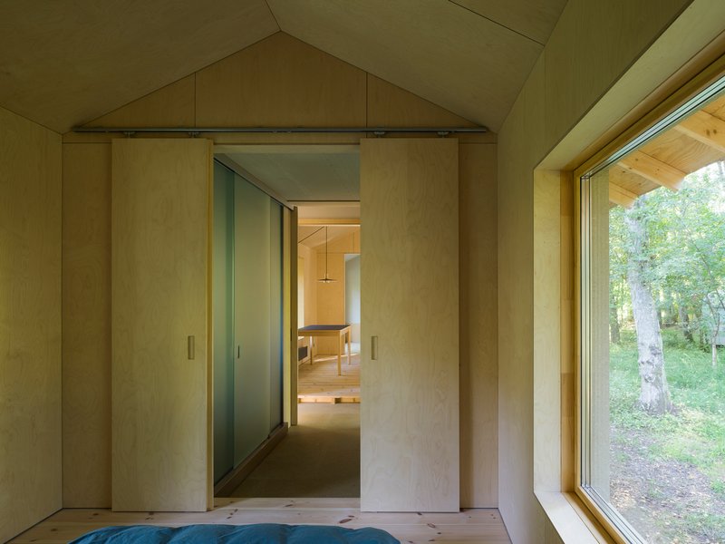 Plywood sliding door opening from a bedroom to a gabled hallway with large window overlooking birch trees