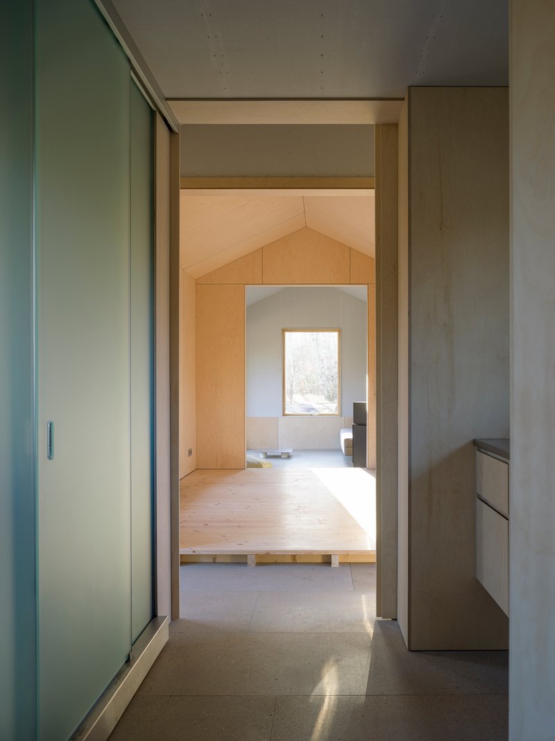 View through doorway showing sequence of plywood-lined rooms with pitched ceilings and natural light