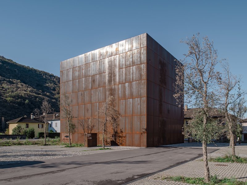 Weathered copper cladding on a cubic volume with perforated panels beside young trees and a hillside