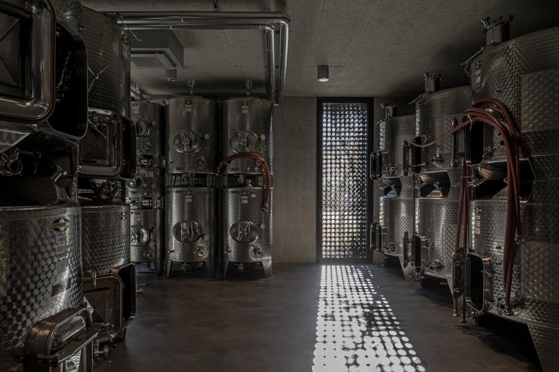Stainless steel fermentation tanks flanking a corridor with dappled light from a perforated wall