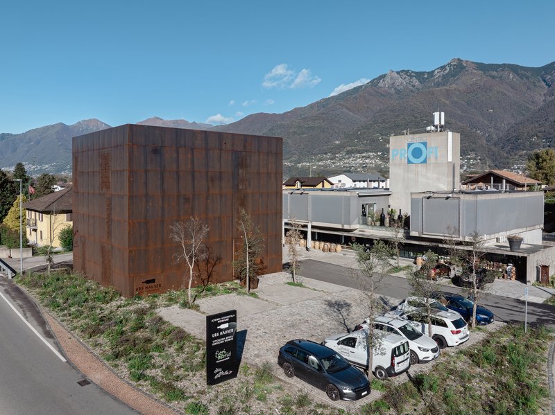 Weathered steel paneled cube adjacent to industrial buildings with mountains in the background