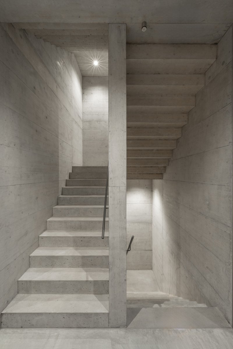 Concrete staircase wrapping around a central column with wall sconces illuminating the board-formed surfaces