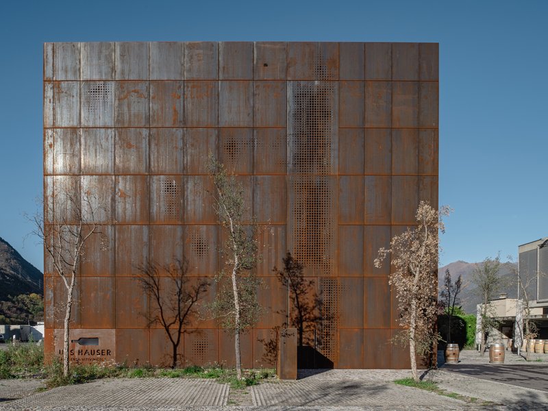 Weathered corten steel facade with perforated panel openings and young trees in front under clear sky