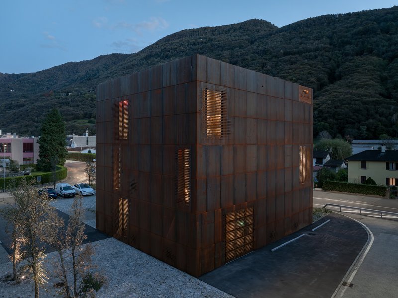 Corten steel cube with illuminated perforated openings at dusk against forested hillside backdrop