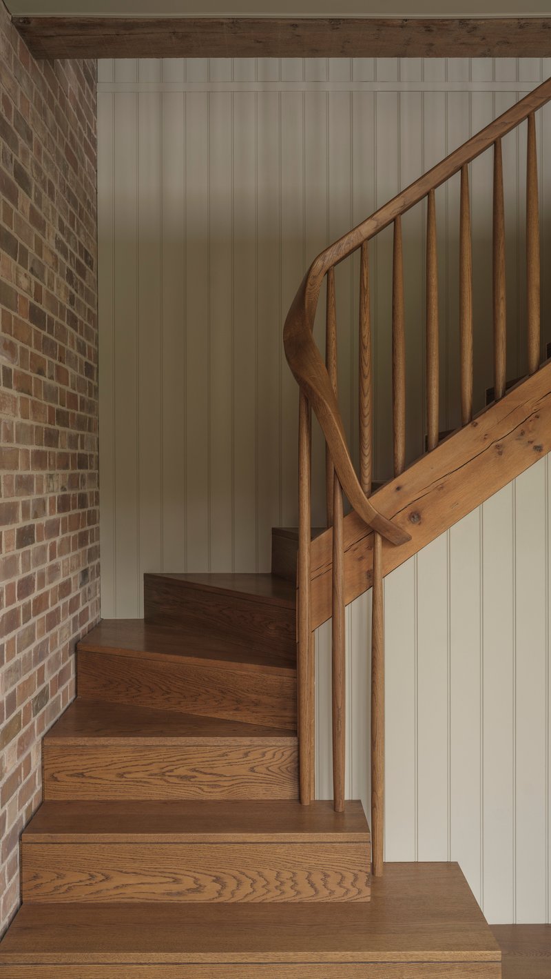 Curved timber staircase rising between brick wall and painted vertical paneling under exposed beam