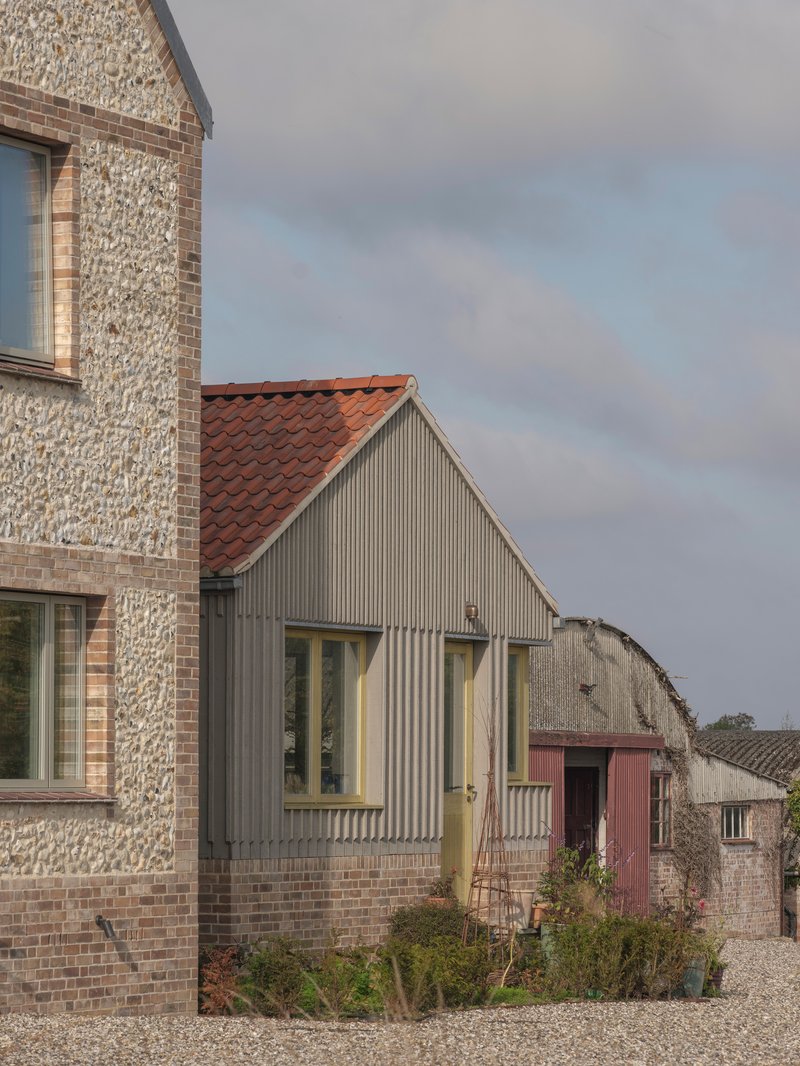 Gable end with vertical timber cladding and terracotta roof beside flint-faced volume and neighboring farm structures