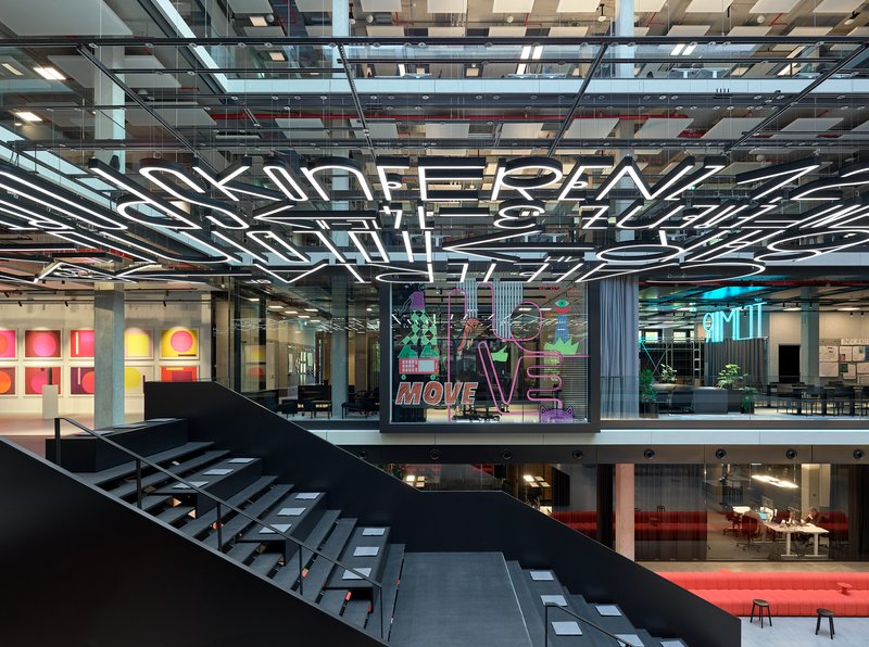 Multi-level atrium with black steel staircase and glazed offices featuring neon signage on upper floors