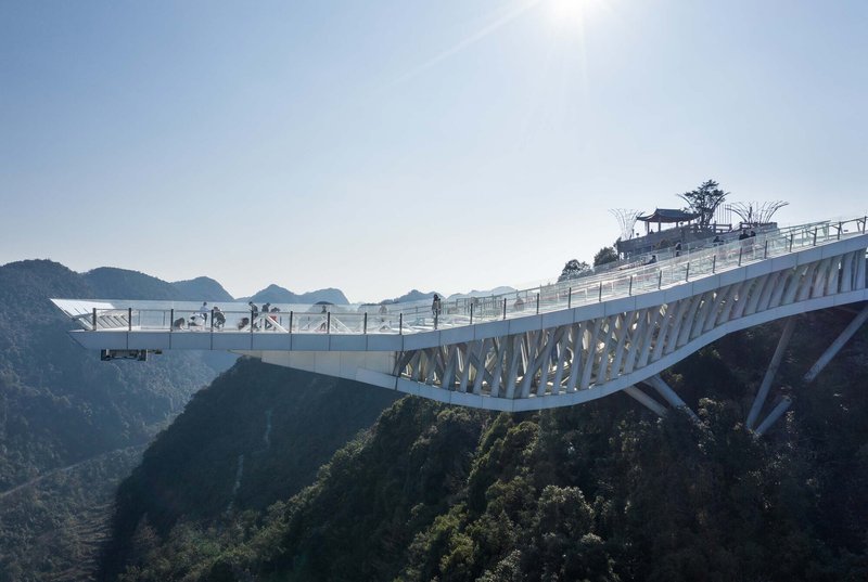 Cantilevered observation deck with diagonal white steel truss extending over forested mountains under clear skies