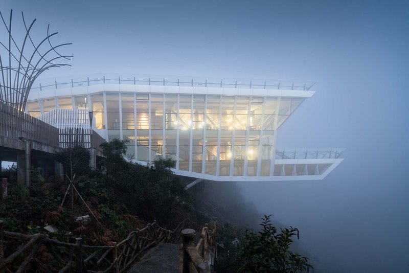 Multi-level glass pavilion with white structural frame glowing at twilight in dense fog