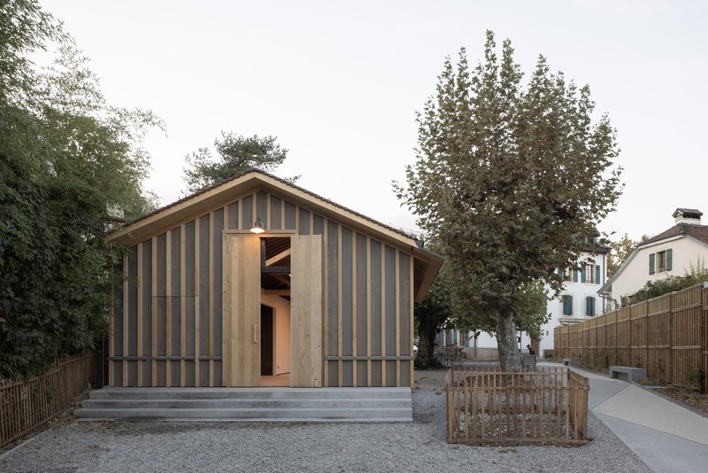Gabled volume with vertical timber and panel cladding facing a gravel courtyard with a central tree