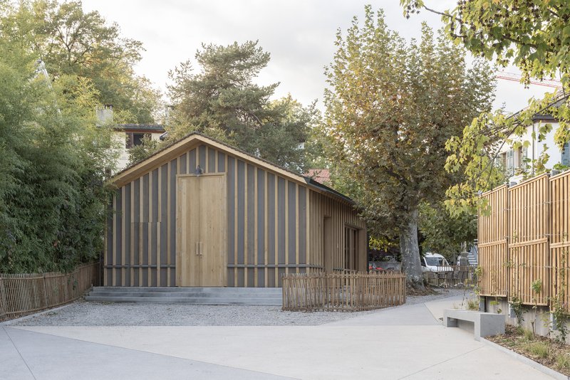 Timber-clad gabled volume with vertical slat facade surrounded by mature trees and gravel courtyard