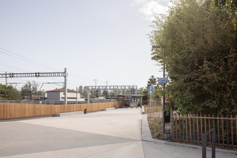 Public plaza with timber-screened walkway leading toward a railway footbridge in overcast daylight
