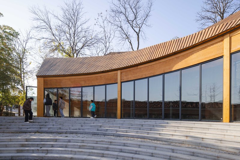 Wide stepped plaza leading to glazed facade beneath a curving corrugated roof with pedestrians ascending
