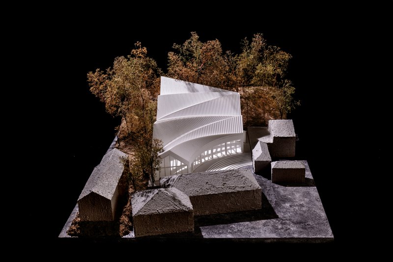 Physical model with ribbed white roof volume positioned among textured concrete blocks and autumn trees