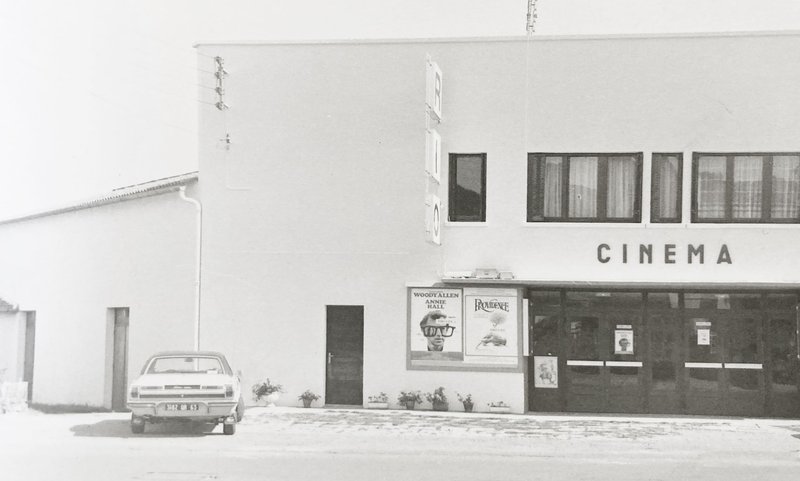 White stucco cinema facade with rectangular windows and a parked car in front