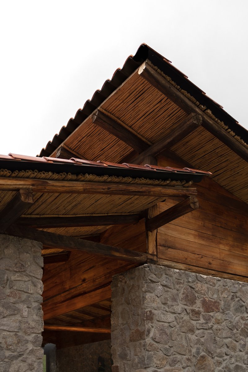 Upward view of timber eaves with exposed rafters meeting a fieldstone corner column under overcast sky