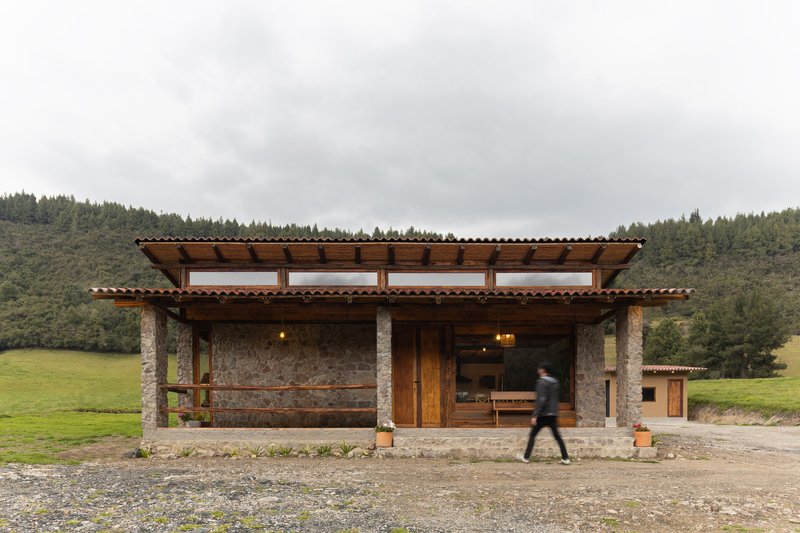 Open pavilion with stone columns and exposed timber roof structure as a figure walks past under overcast skies