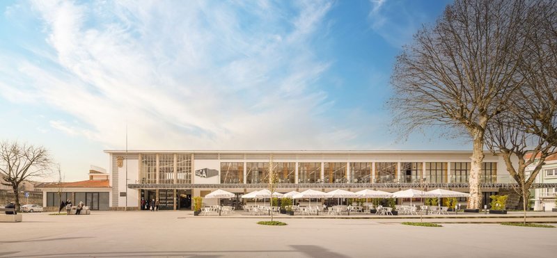 Frontal elevation of the glass-walled market hall facade with bare trees framing the entrance plaza