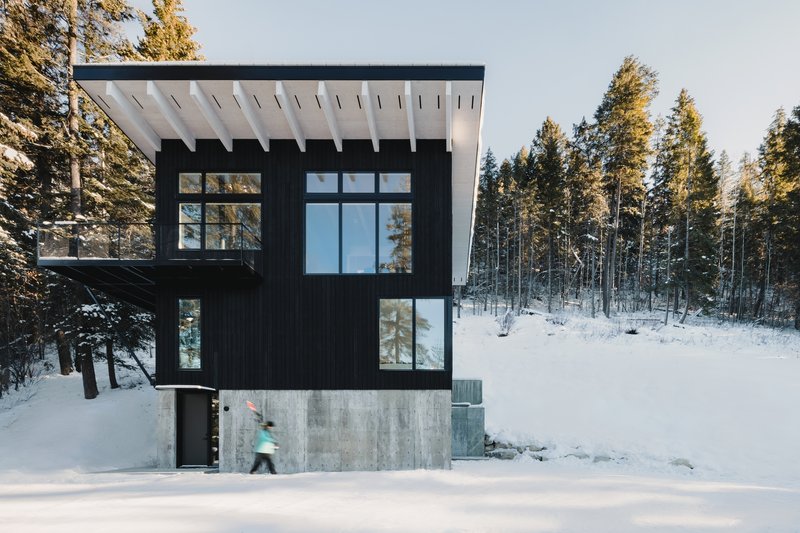 Black timber clad volume with gridded windows on a concrete base in a snow covered forest with a figure passing by