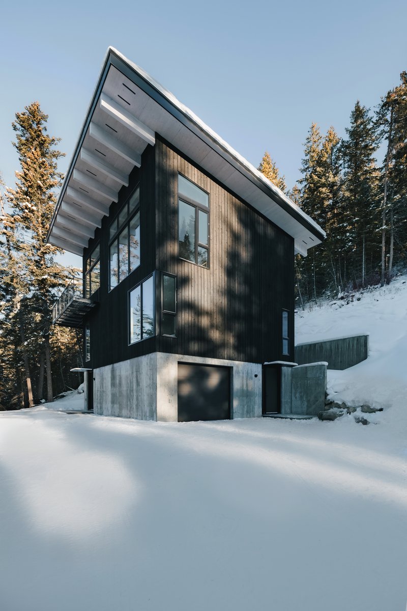 Two-storey facade showing dark cladding on exposed concrete plinth surrounded by deep snow and pine trees in afternoon light