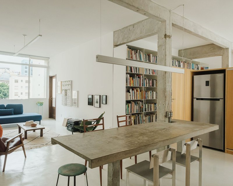 Open living and dining area with exposed concrete frame, bookshelf wall, and light from a large window