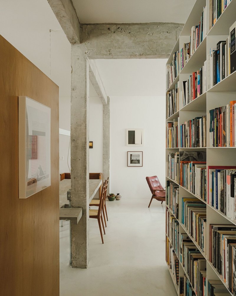 Corridor view past concrete columns toward white bookshelves and a red leather chair