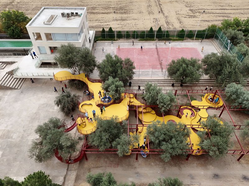 Aerial view of the yellow curving playground structure with red railings among olive trees and a tennis court