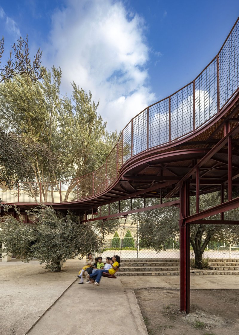 Curved canopy with mesh guardrails sheltering steps and seating where children gather among the olive trees
