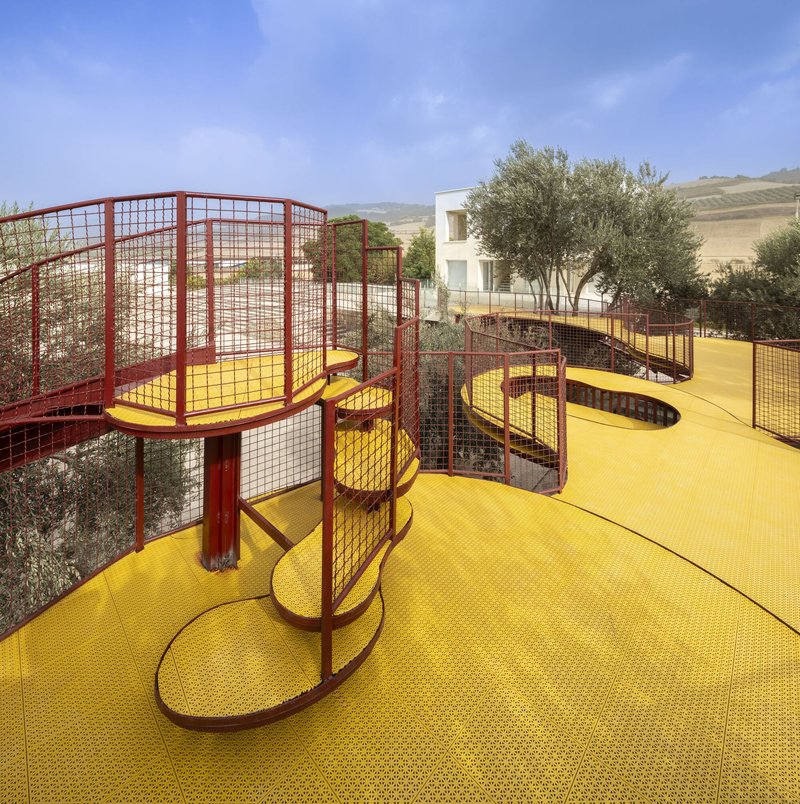 Elevated yellow walkway with red mesh railings winding between olive trees under a clear blue sky