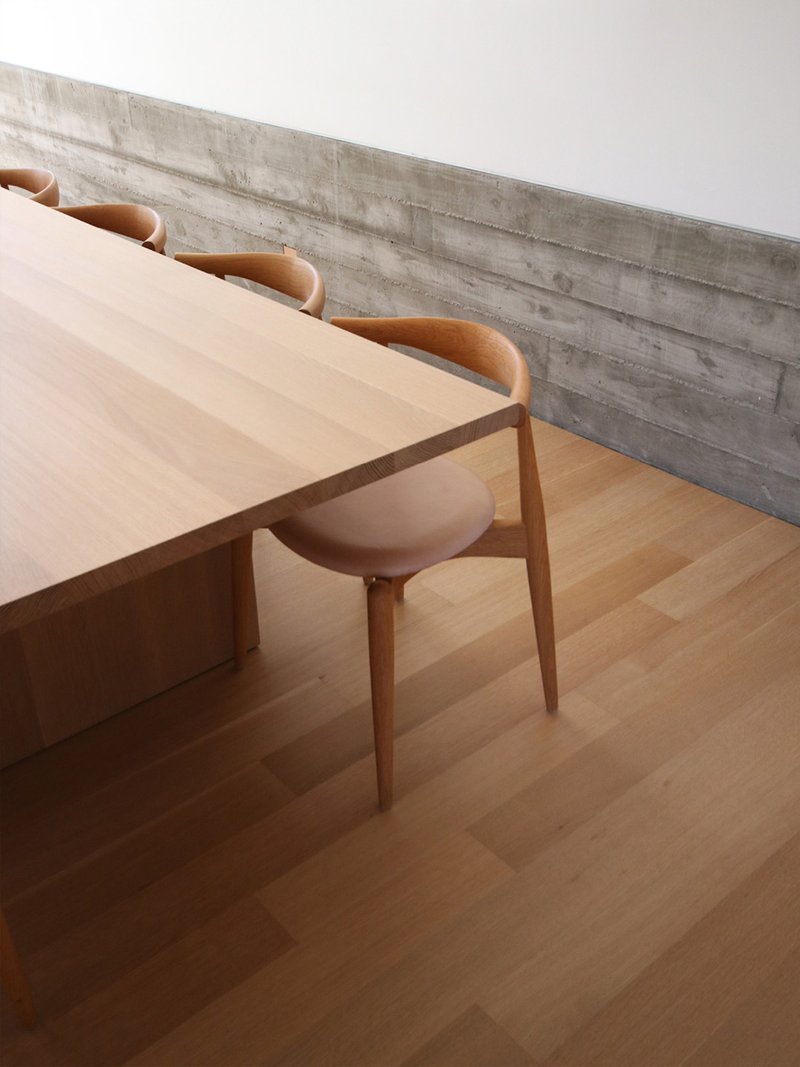 Light timber dining table and chairs against a board-formed concrete wall in soft natural light