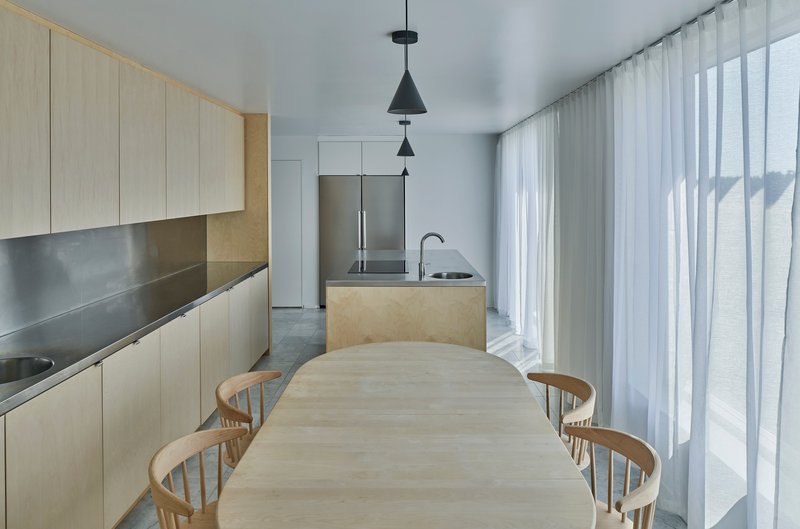 Open kitchen and dining area with pale timber cabinetry, stainless steel countertops, and sheer curtains filtering daylight