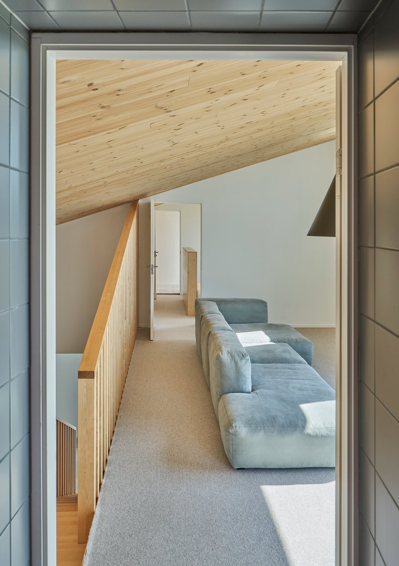 View through a doorway toward a hallway with angled plywood ceiling and timber slatted balustrade catching afternoon sun