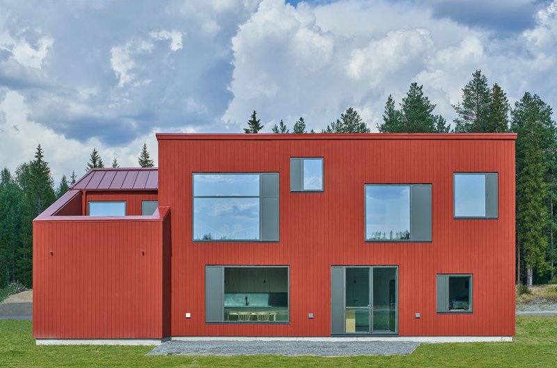 Facade with asymmetrically placed windows in the red vertical cladding overlooking a gravel yard