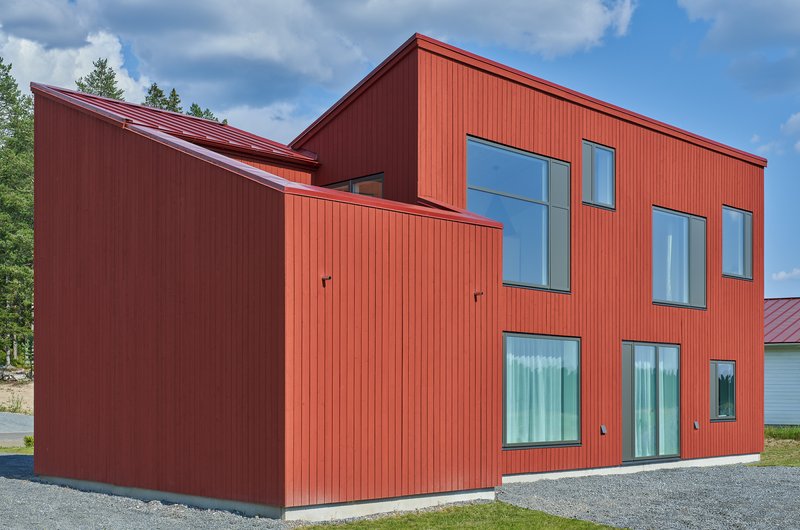 Corner view showing the red ribbed siding and projecting volumes beneath white clouds