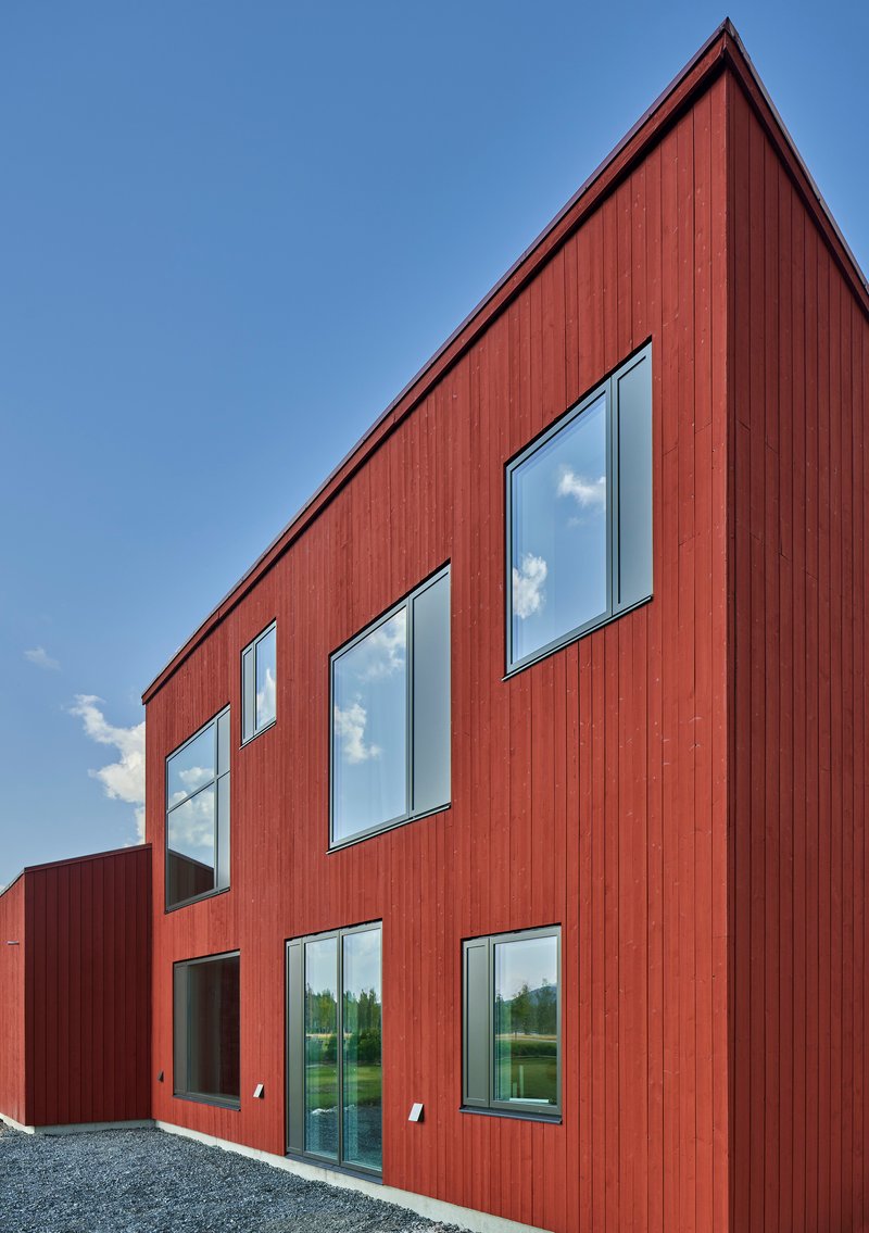 Red vertical wood siding facade with six punched windows reflecting clouds against a clear blue sky