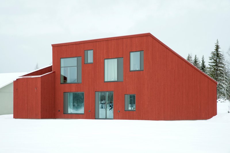 Two-story red cladded volume with asymmetrical windows sitting in deep snow beside a distant structure
