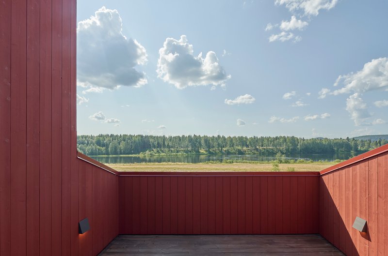 Roof terrace enclosed by red wood paneling framing a lake and forest under a cloudy summer sky