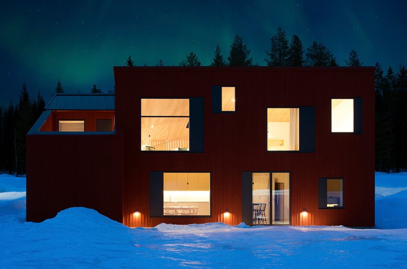 Illuminated windows at dusk reveal interior spaces while three wall-mounted lights glow against snowbanks